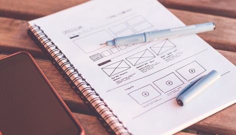 Ein iPhone, ein Notizblock und Faserstift liegen auf. Darauf ist eine Skizze eines Wireframes.