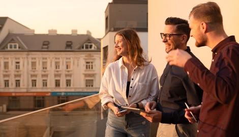 Drei Mitarbeiter stehen auf einer Terrasse im Sonnenuntergang und unterhalten sich lachend.