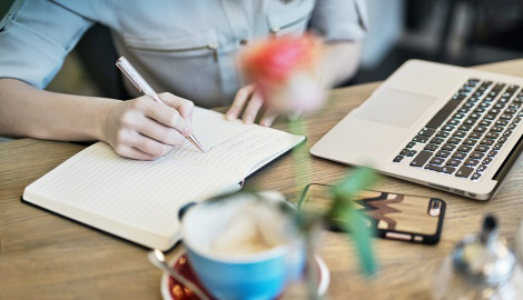 Frau vor einem Macbook schreibt auf einem Holztisch in ein Notizbuch