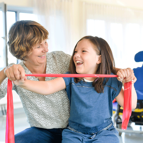 Betreuerin übt mit Kind mit einem Gymnastikband. Dahinter ist ein Rollstuhl zu sehen