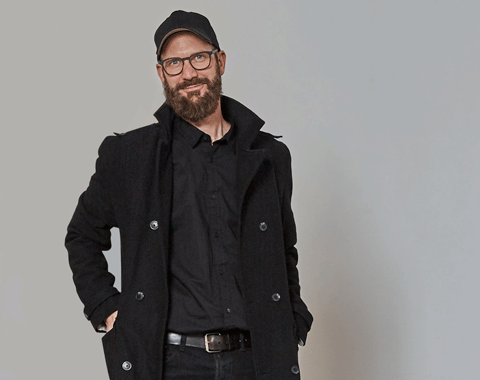Ein Mann mit Bart, Brille, schwarzem Mantel, Hemd, Hose, und Kappe l&auml;chelt in die Kamera.