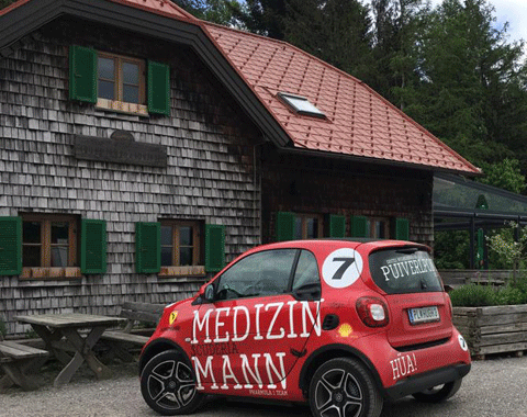 Ein roter Smart mit der wei&szlig;en Aufschrift MEDIZINMANN steht vor einem Holzhaus.