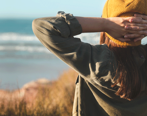 Eine Frau mit Haube, von hinten fotografiert, blickt an einen Strand und hat dabei die H&auml;nde hinter dem Kopf verschr&auml;nkt.