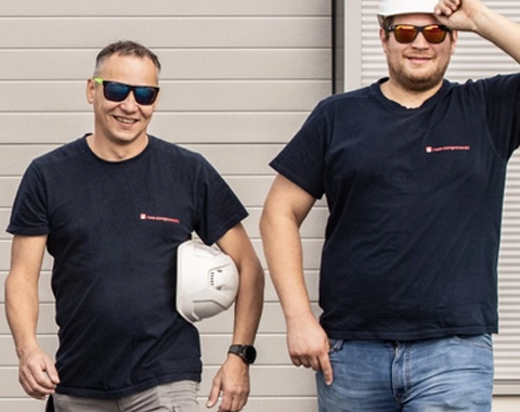 Zwei M&auml;nner gehen l&auml;chelnd mit wei&szlig;en Bauhelmen mit viel Elan auf die Kamera zu.