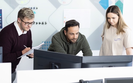 2 Männer und eine Frau stehen vorgebeugt vor einem PC und blicken konzentriert in den Monitor.