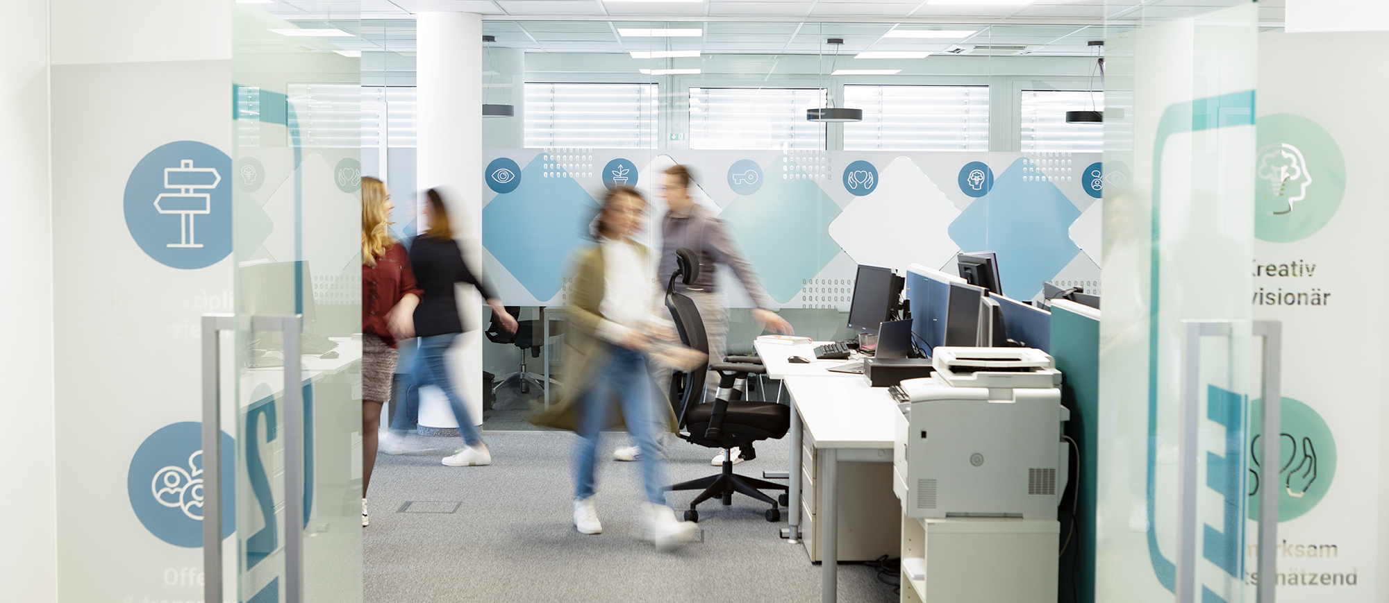 Blick ins Q2E-Büro, man sieht Schreibtische und vier verschwommene Personen durch das Bild laufen