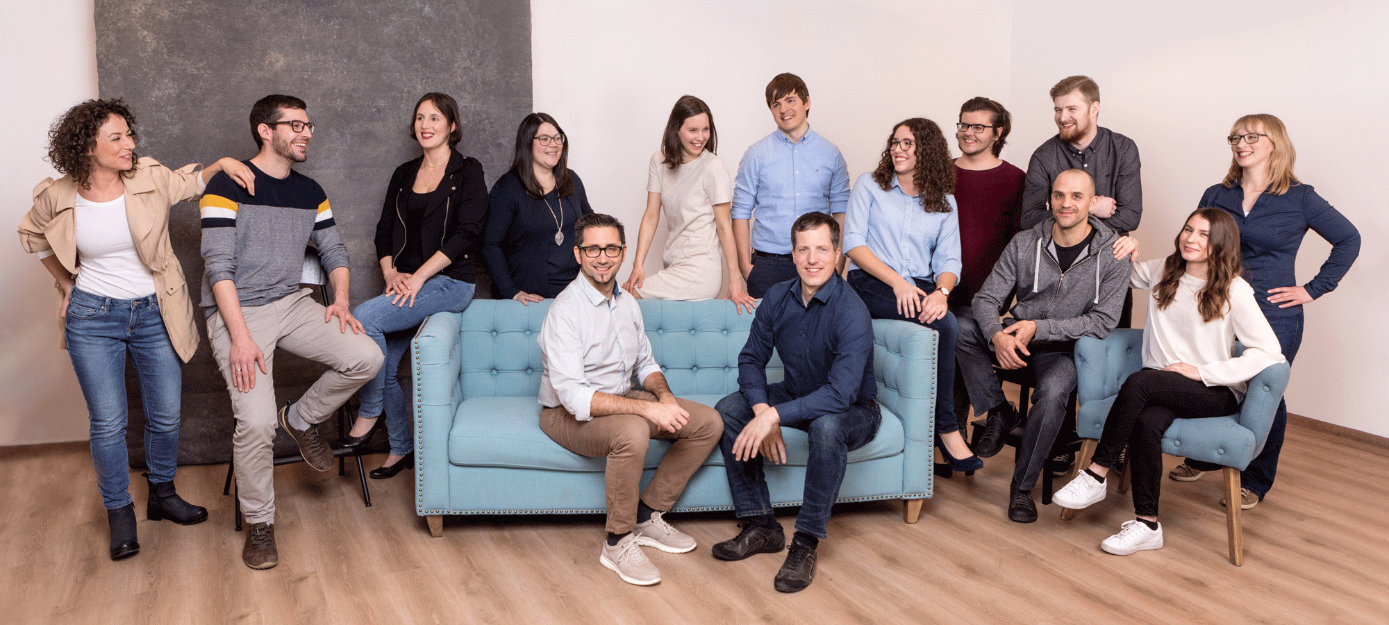 14 Personen stehen teilweise, sitzen teilweise auf einer Couch und lachen in die Kamera.