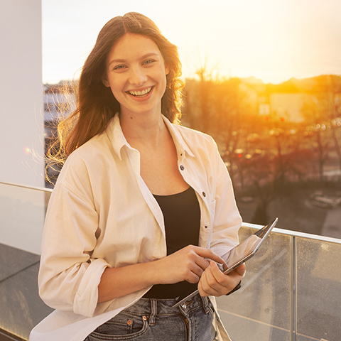 Eine junge Frau in T-Shirt und Hemd steht mit einem Tablet auf einem Balkon im Sonnenuntergang.
