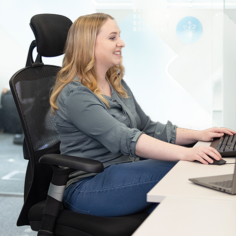 Eine blonde Frau sitzt an einem Schreibtisch und blickt Richtung Monitor