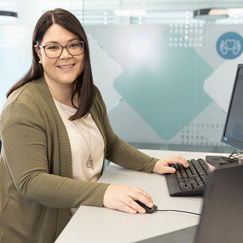 Eine dunkelhaarige Frau mit Brille steht an einem Schreibtisch und lacht in die Kamera.
