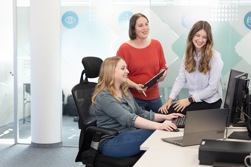 3 Frauen sitzen bzw. stehen vor einem PC und plaudern fröhlich.