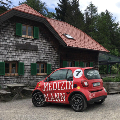 Ein roter Smart mit der weißen Aufschrift MEDIZINMANN steht vor einem Holzhaus.