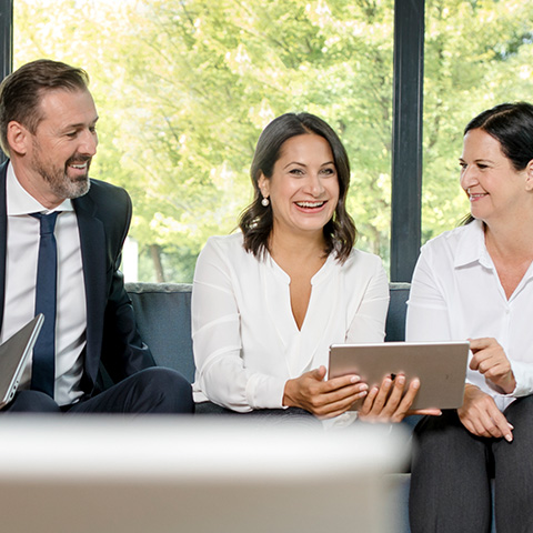 Ein Mann und zwei Frauen in förmlicher Kleidung sitzen mit einem Tablet nebeneinander und lachen.