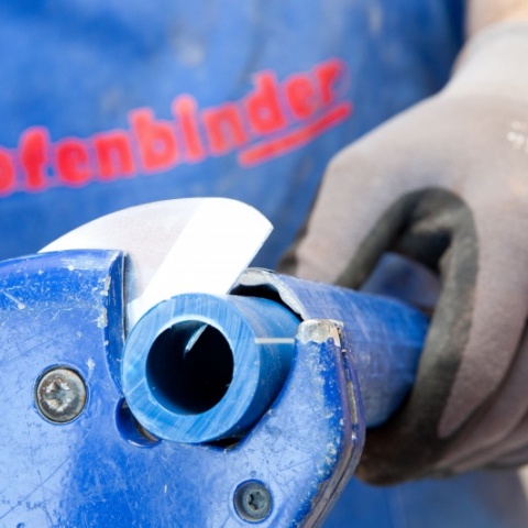 Ein blaues Rohr wird mit einem Werkzeug von einem Ofenbinder-Mitarbeiter abgeschnitten.