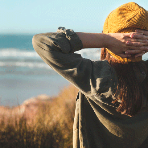 Eine Frau mit Haube, von hinten fotografiert, blickt an einen Strand und hat dabei die Hände hinter dem Kopf verschränkt.